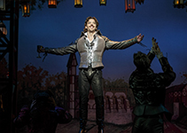 Christian Borle wearing a mordern fashionable leather Elizabethan styled jacket, pants, and large codpiece and has his arms osutsretched as he sings.